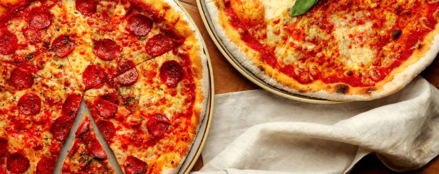 close-up view of two artisan pizzas served on plates next to each other on a wooden surface with a white linen napkin; one is a pepperoni pizza and the other is a classic margherita with fresh basil