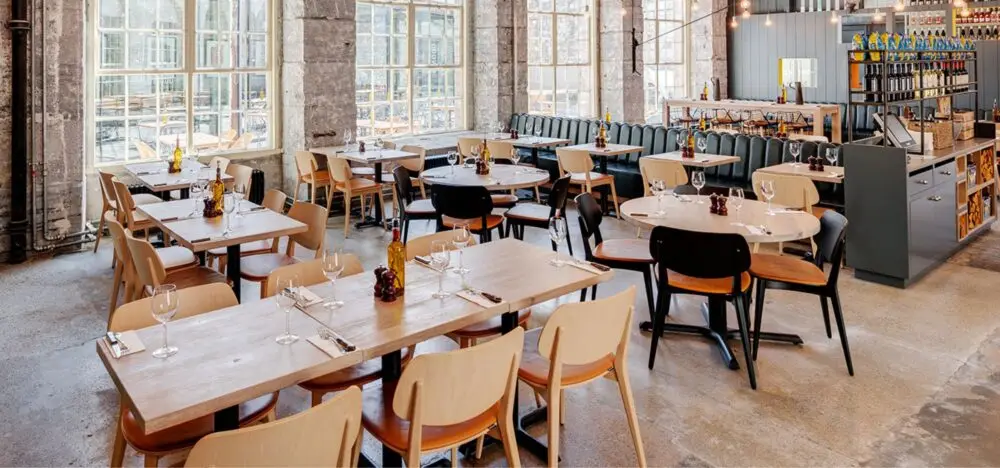 Interior dining area of Wildwood at Royal William Yard in Plymouth, featuring a modern, industrial design with exposed stone walls, large bright windows, light-wood tables, and tan upholstered chairs