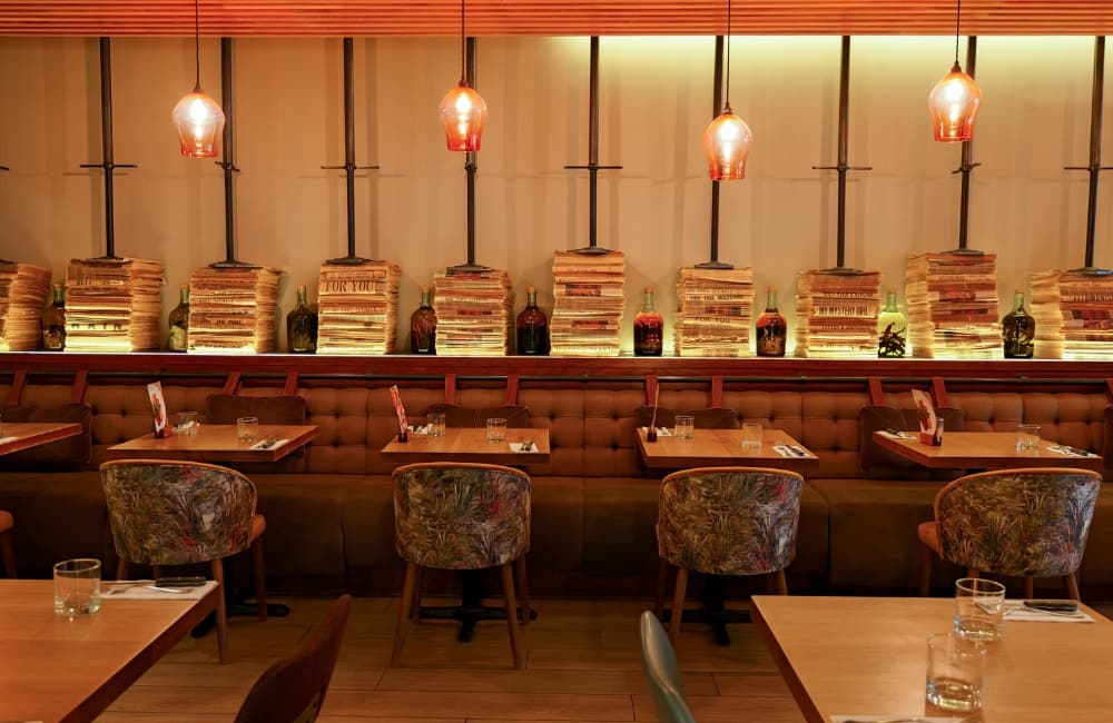 Interior dining room at Wildwood Covent Garden featuring wooden tables paired with patterned tub chairs, a long button-tufted banquette bench, glowing orange pendant lights, and decorative stacks of vintage newspapers alongside olive oil bottles.